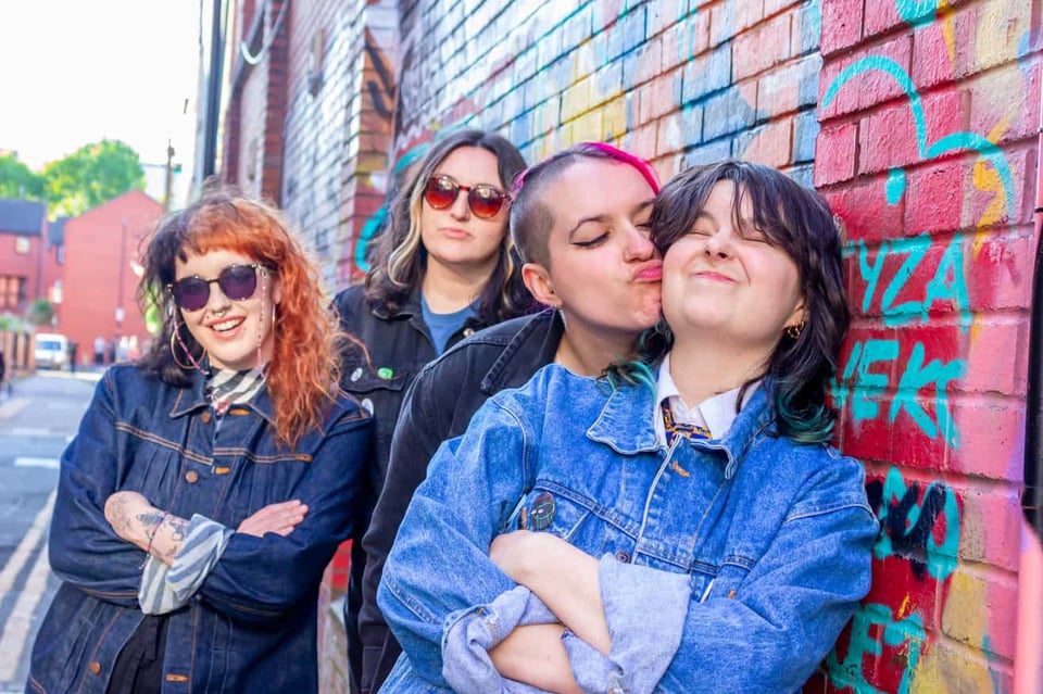 Four young people standing next to a wall covered with colorful graffiti. They are all wearing jeans jackets. One of them is smushing her face against another one's face and they are both squinting and making faces.