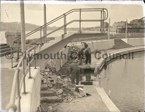 Several workmen working on a hole that has been blasted in the inner seaward curve of the Lido pool. A short concrete diving platform sticks out above them.