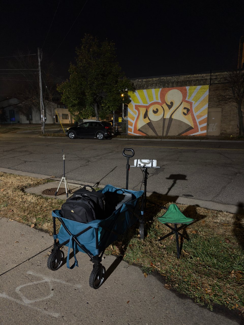 Photo of a night scene with a mural that says love across the street from my projection mapping set up. Set up includes a blue soft sided wagon with 2 backpacks, a green tripod stool, my projector on a projector stand, and a tripod for recording.