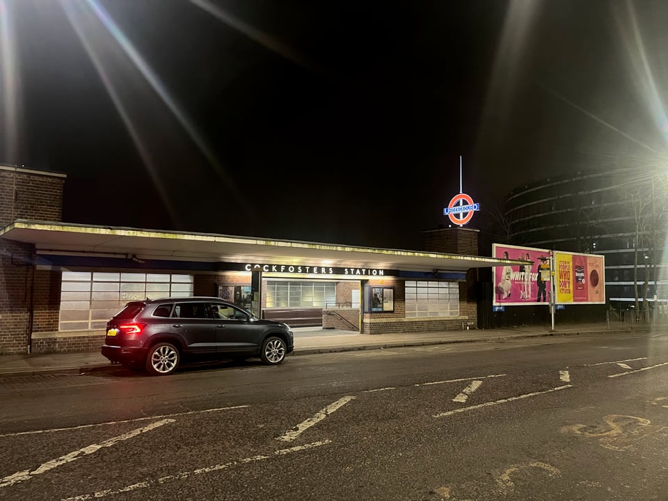 Cockfosters Station at night. I'm standing across the road and looking towards it; the night sky is pitch black and the station is lit up. It looks warm and cosy inside! Behind the main building is an illuminated Tube roundel raised high in the sky.
