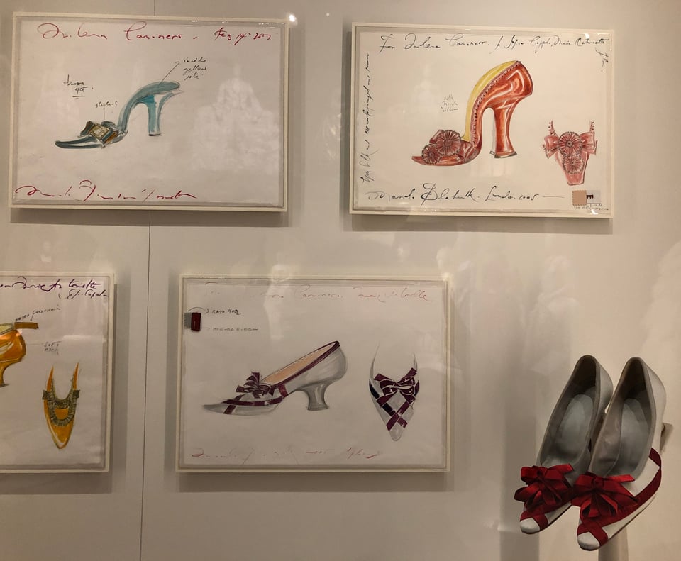 Three and a half framed designs for brightly coloured shoes, and a pair of shoes in the same style, in an exhibition case. Each design contains a profile view of a very high-heeled woman’s shoe, a front view of the toe, and some text. The pair of shoes is a whitish-grey, with deep red ribbons and a large rosette on the toe and front of the foot.
