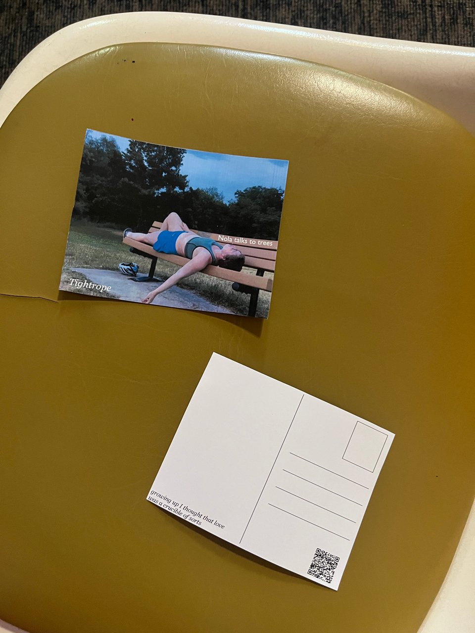 Tightrope postcards, front and back, on a retro dark yellow vinyl chair. The front side is Nola lying on a bench against trees and cloudy blue sky; the back is a classic postcard layout with the quote "growing up I thought that love was a crucible of sorts" and a QR code.