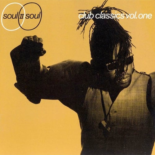 A yellow-tinted black and white photo of Soul II Soul bandleader Jazzie B, from the waist up. He's a Black man wearing a vest and a necklace with Africa on it. He has dreadlocks, and they're mostly tied up out of is face save for a few strands falling across his face. He is the anti-Don Henley. He appears to be pumping his fist. He looks to the right with a blank expression.