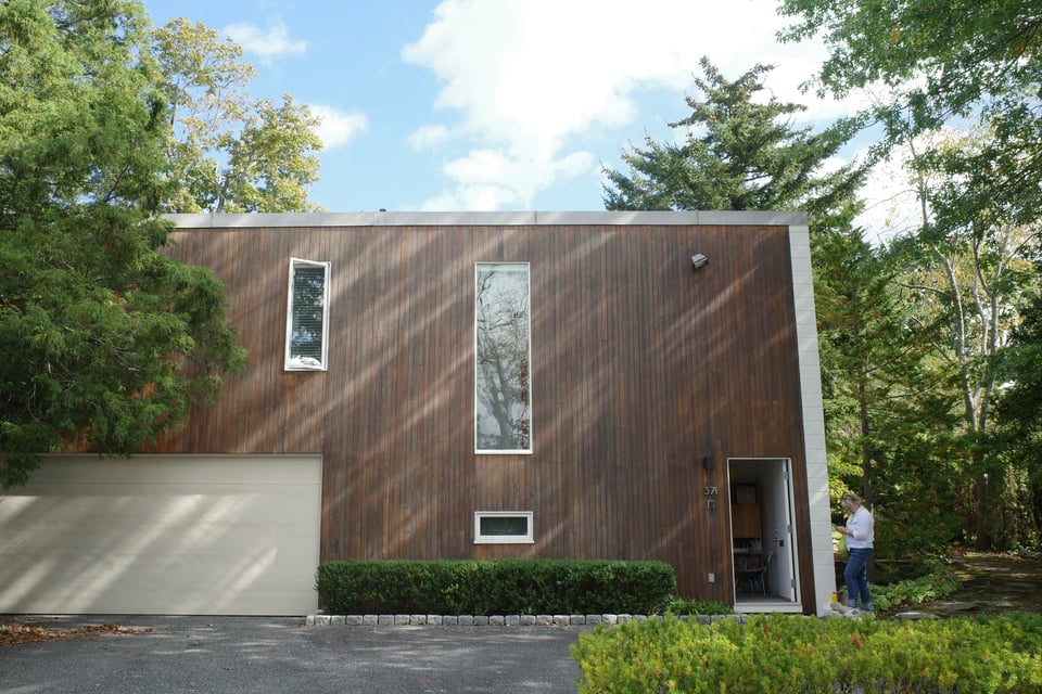 Contemporary house clad in vertical warm wood siding with tall narrow windows, a light garage door, and dense trees surrounding the site.
