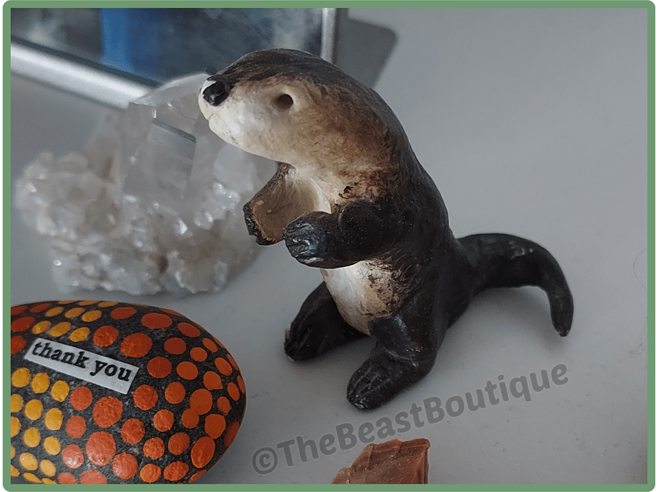 a photograph of a roughly sculpted otter figurine standing on its back legs. It sits next to a painted rock and a small cluster of quartz crystals.