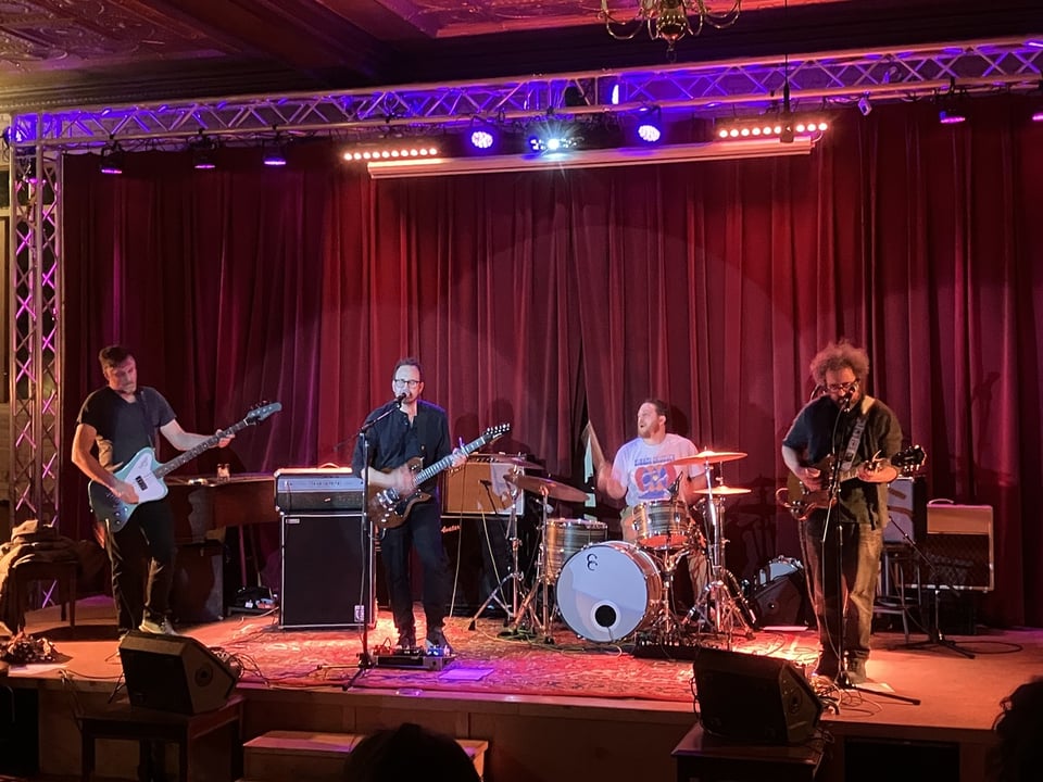 A bassist, guitarist, drummer and second guitarist perform onstage in front of a red curtain.