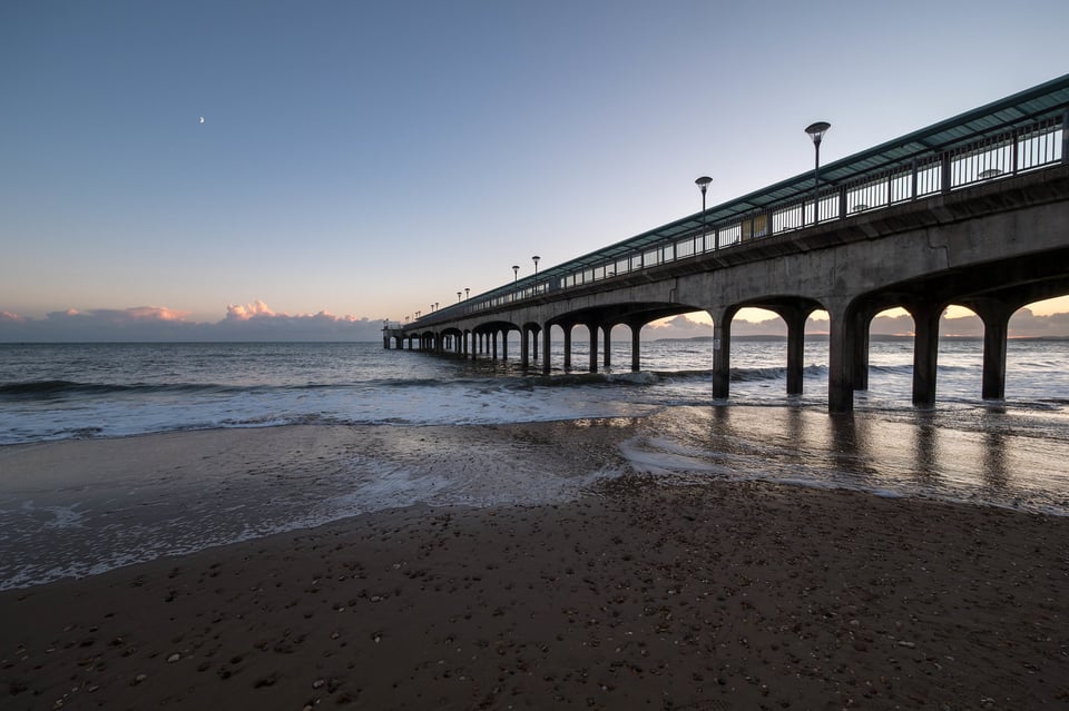 A pier stretching out into the sea at sunset. Instead of iron legs, it has concrete ones, reaching up to gently curved arches. A simple Y shaped shelter runs the entire length of the pier.