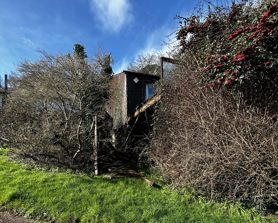 Hugely overgrown bushes. Lurking within is the remains of the first pair of lodges. The privacy screen is missing but the cantilevered balcony is still there. The wooden cladding is fading and thick with ivy.