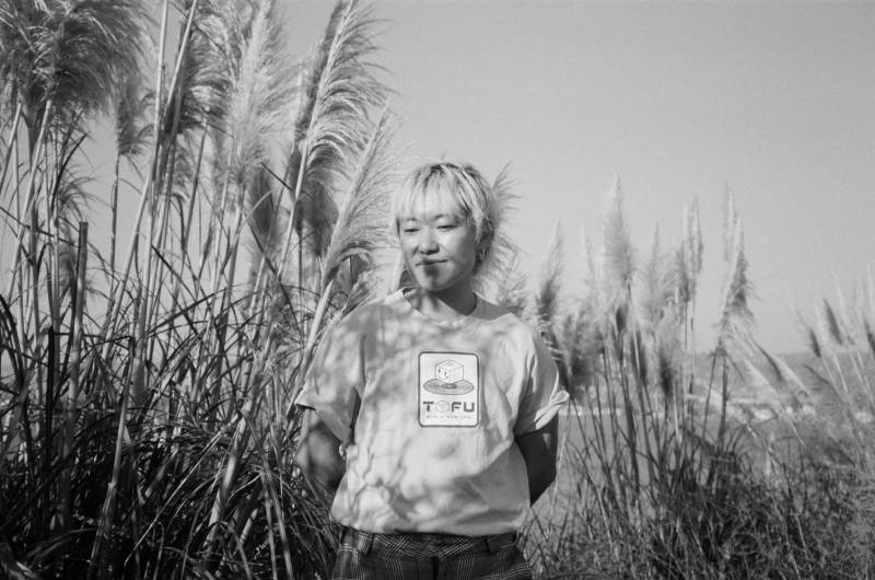A blonde gremlin stands in front of a few bushels of tall, feathery pampas grass. A black and white photo taken by Menat el Attma at the Albany Bulb.