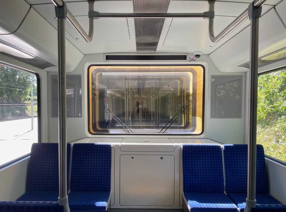 An S-bahn (local city train) compartment's view of a large glass at the end looking into the next compartment. The bright yellow outer paint bathed in sunlight from the next compartment appears to be some sort of a ring light. Four, blue checkered seats lay empty with stainless steel holding bars forming clean geometric shapes visible in the foreground.