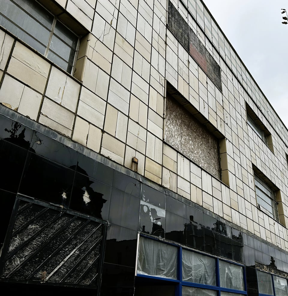 A three stoey elevation. The ground floor has retail units that have their windows obscured and is clad in black vitrine tiles. Several of these are missing and another has clearly been water damaged. The upper storeys are clad in cream faience tiles in a grid pattern. Some of these are cracked or discoloured. Some of the windows are literally boarded up with chipboard.