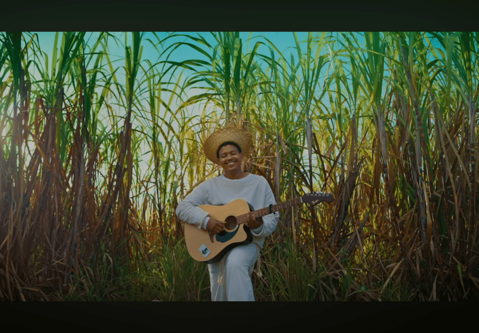 A man in all white and a straw hat, holding with a cutaway acoustic guitar, smiles as he introduces Bad Bunny's Super Bowl halftime show.