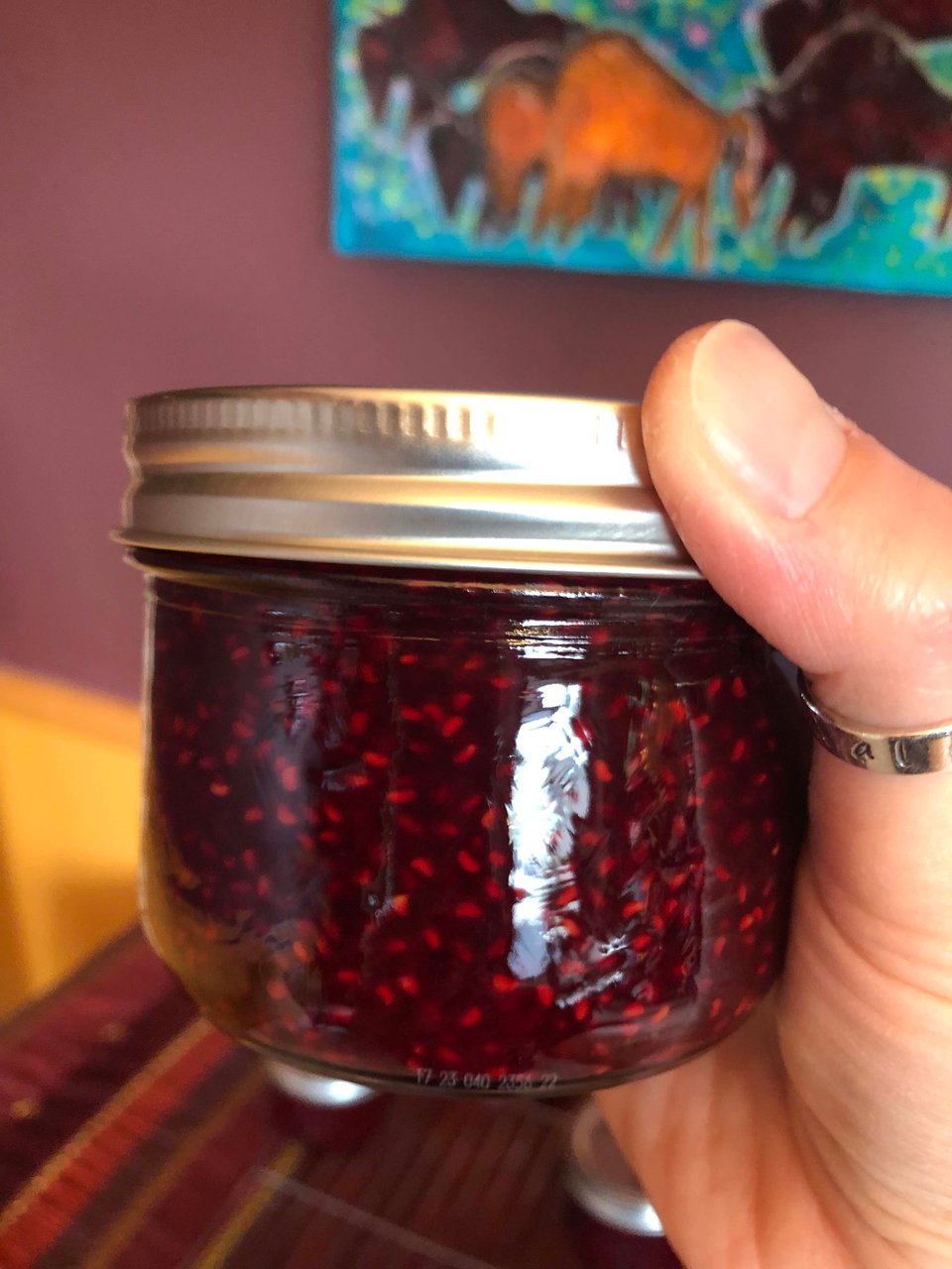 A pale hand (mine) wearing a silver thumb rung holds a squat jar. Inside is dark red jam with seeds. In the background on the purple wall is part of the painting, Coming Home by Dawn-Marie Marchand, showing a number of gouache bison in light and dark brown, on bright aqua grass dotted with pink flowers