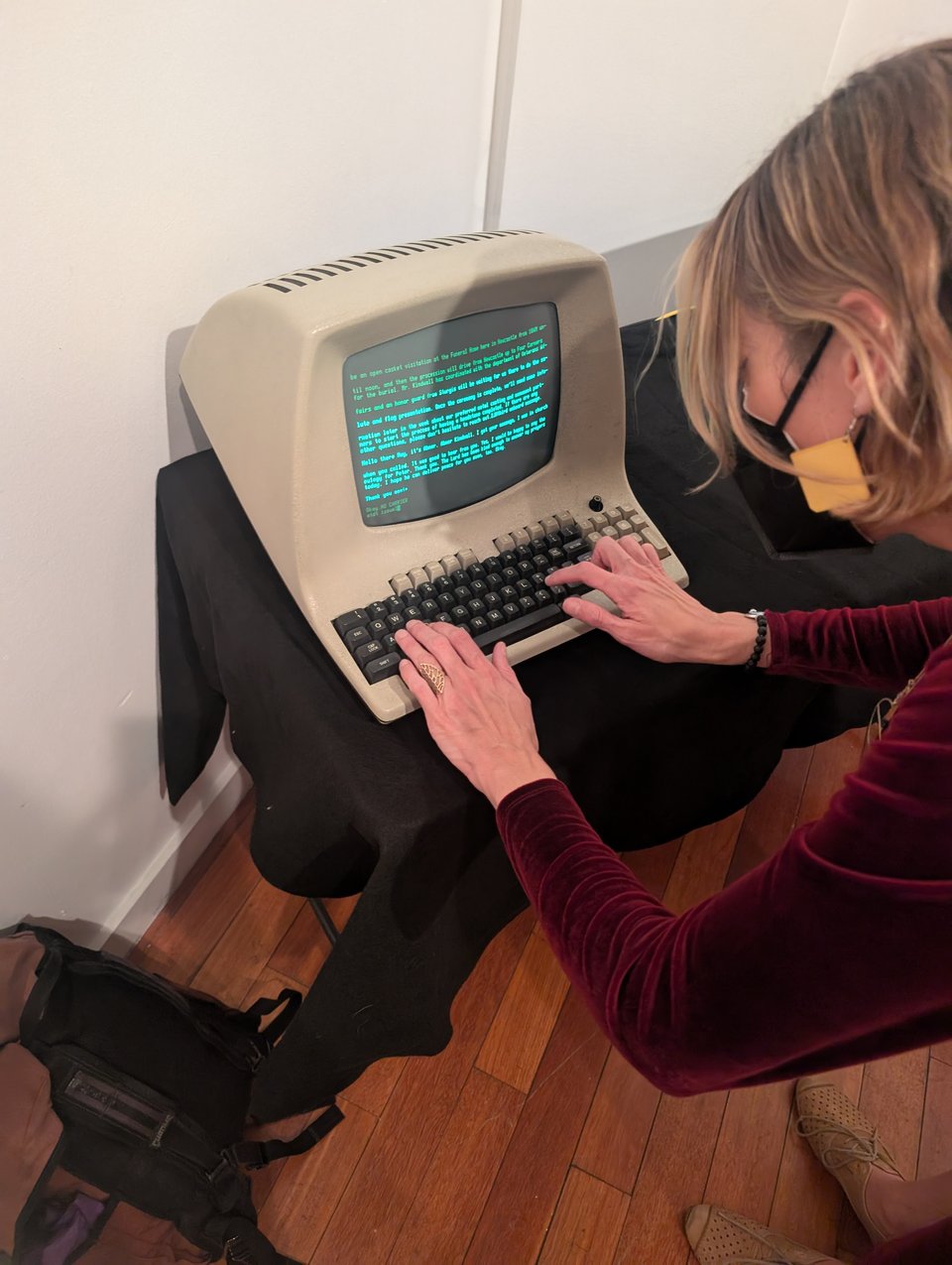 A long-haired person wearing a mask and a red top types at a very old computer terminal with green lettering