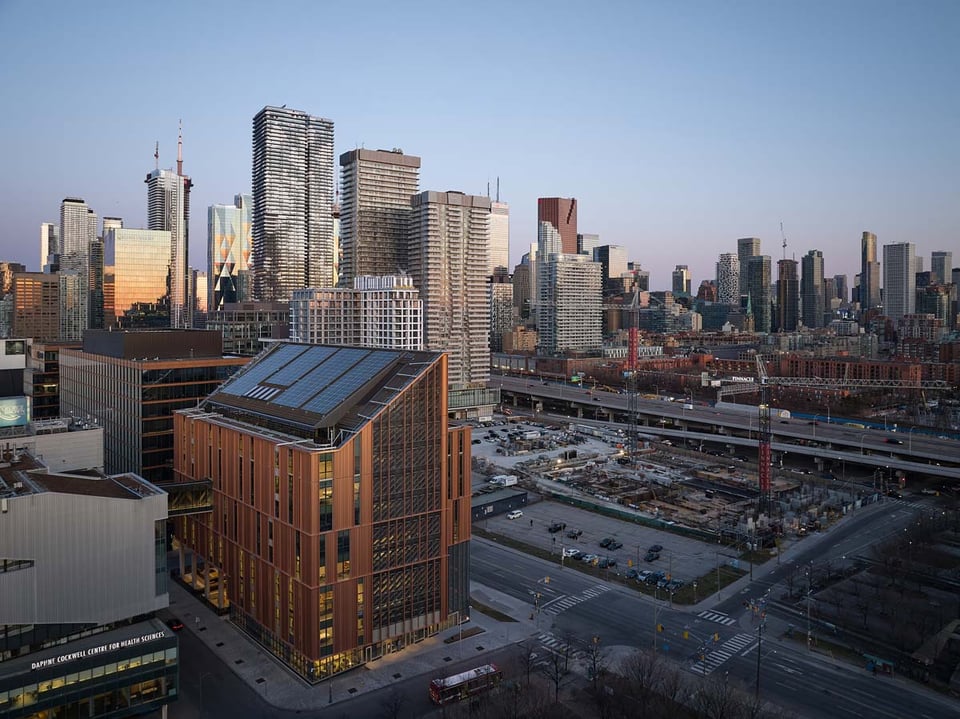 A cityscape at dusk showing a modern downtown skyline with glass high-rises and office towers reflecting the fading sunlight. In the foreground stands a tall, angular building with solar panels on its roof, made of wood that's a medium-dark, walnut-brown
