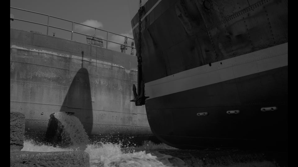 A black and white photo of the front of a large ship taking up the right half of the screen. It is sitting in a dry dock and we see it's hull. On the right is a large wall with a small hole where water is gushing out of as the dry dock slowly fills up. In the top left of the image you see a few puffy clouds and clear but desaturated sky.