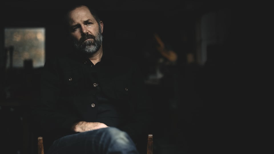 A man with a full salt-and-pepper beard sits in a chair in a dimly-lit room, with his right leg crossed over his left.