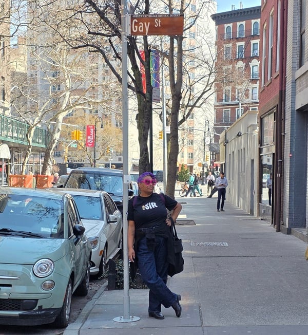 Photo of Patricia wearing a black t-shirt with Peppermint Patty on it that says “SIR.” Patricia is standing under a street sign that says “Gay Street.”