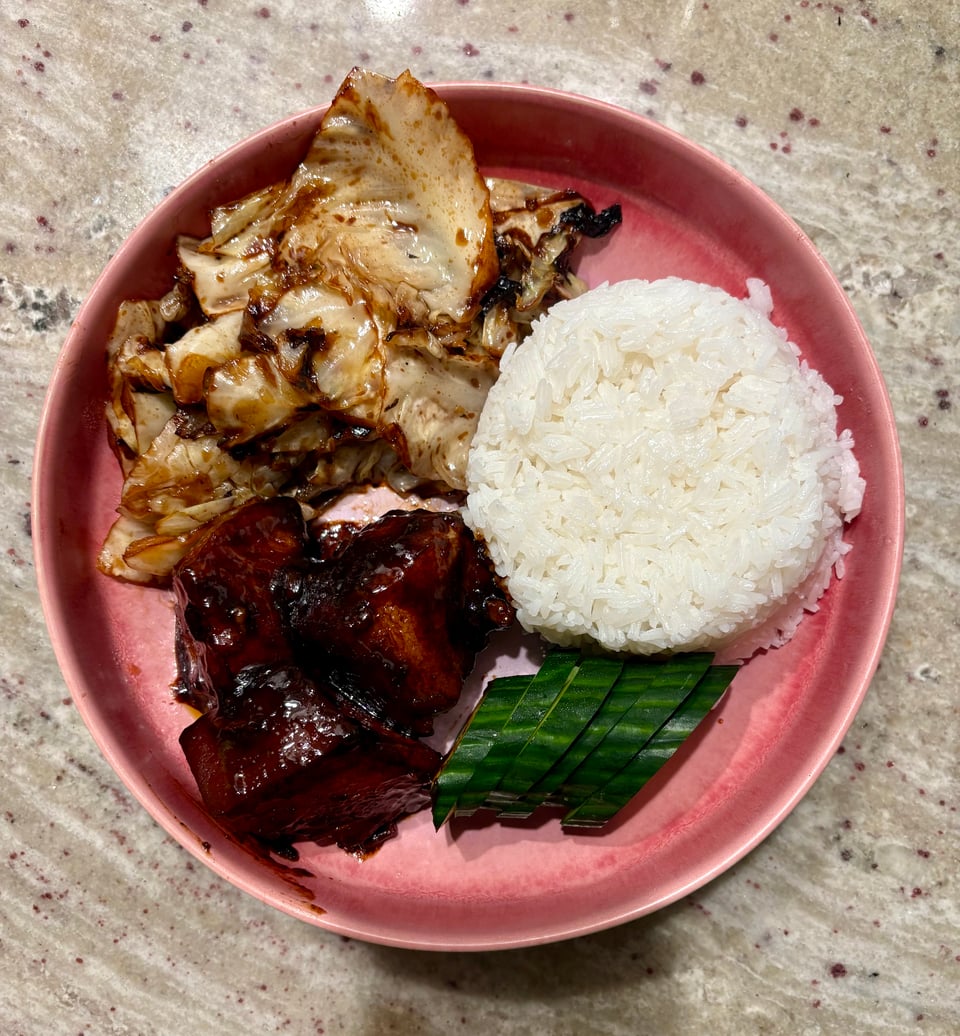 A pink plate of braised pork belly, sliced cucumber, jasmine rice, and simple stir-fried cabbage.