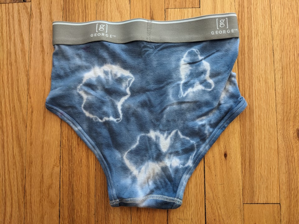 A pair of tie-dyed briefs, blue with white bulls-eyes and a gray waistband, against a hardwood floor.