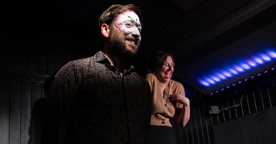 Gael and Mash on stage with makeup, Mash in the background as a clown and Gael in the foreground as a clock. Both are smiling.
