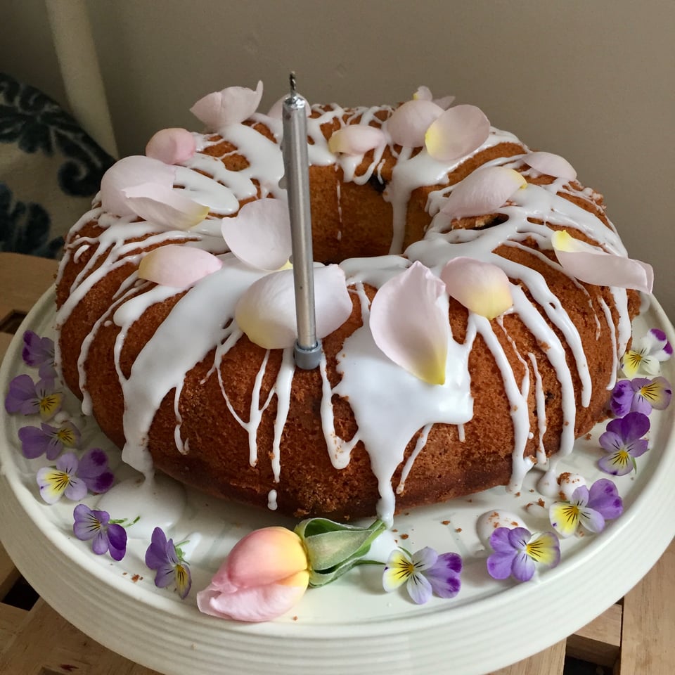 A round bundt cake is drizzled with white icing and scattered with pale pink rose petals and surrounded by purple and yellow pansies. Image by Rowan Ambrose