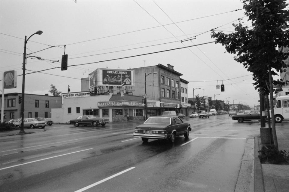 Street scene of an intersection. You can see a restaurant in the midground. There are trees planted all along the sidewalk.