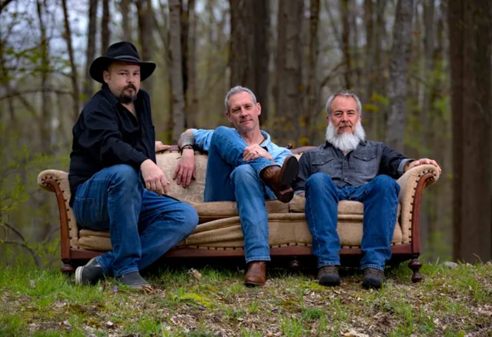 Three men in bluejeans sit on a battered couch in the woods.