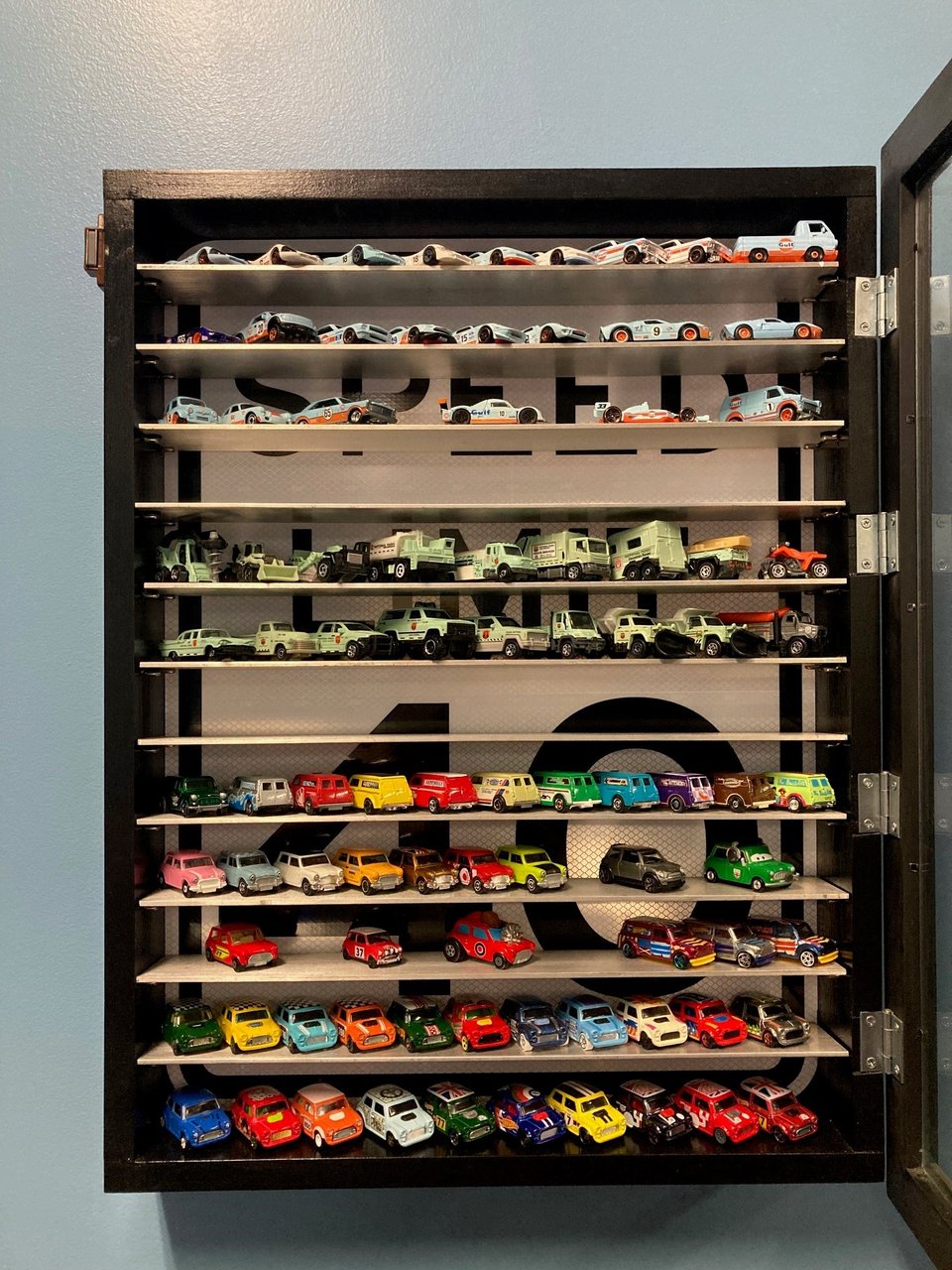 A display case featuring numerous diecast vehicles on aluminum shelves. The background of the display case is a speed limit sign.