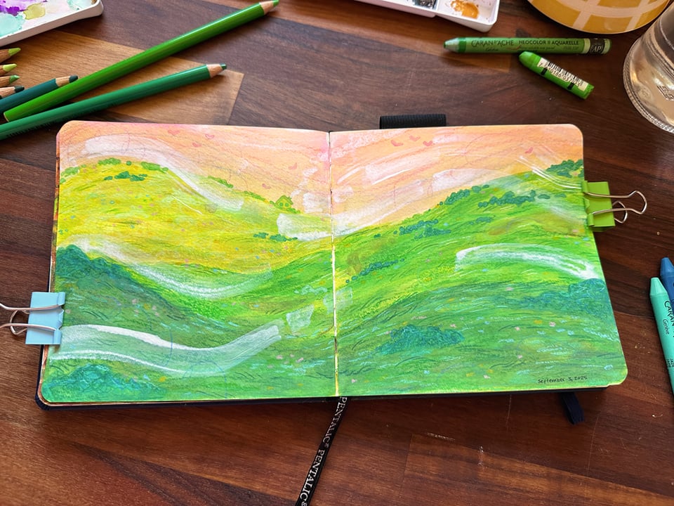 A messy, experimental spread in a small sketchbook showing a landscape created with mixed media. The sky is a sherbert hue of orange, contrasted against the vibrant greens of the rolling hills in the foreground. In the distance, a single house pipes smoke from its chimney, and gusts of wind are drawn across the landscape. The effect is a peaceful enough one.