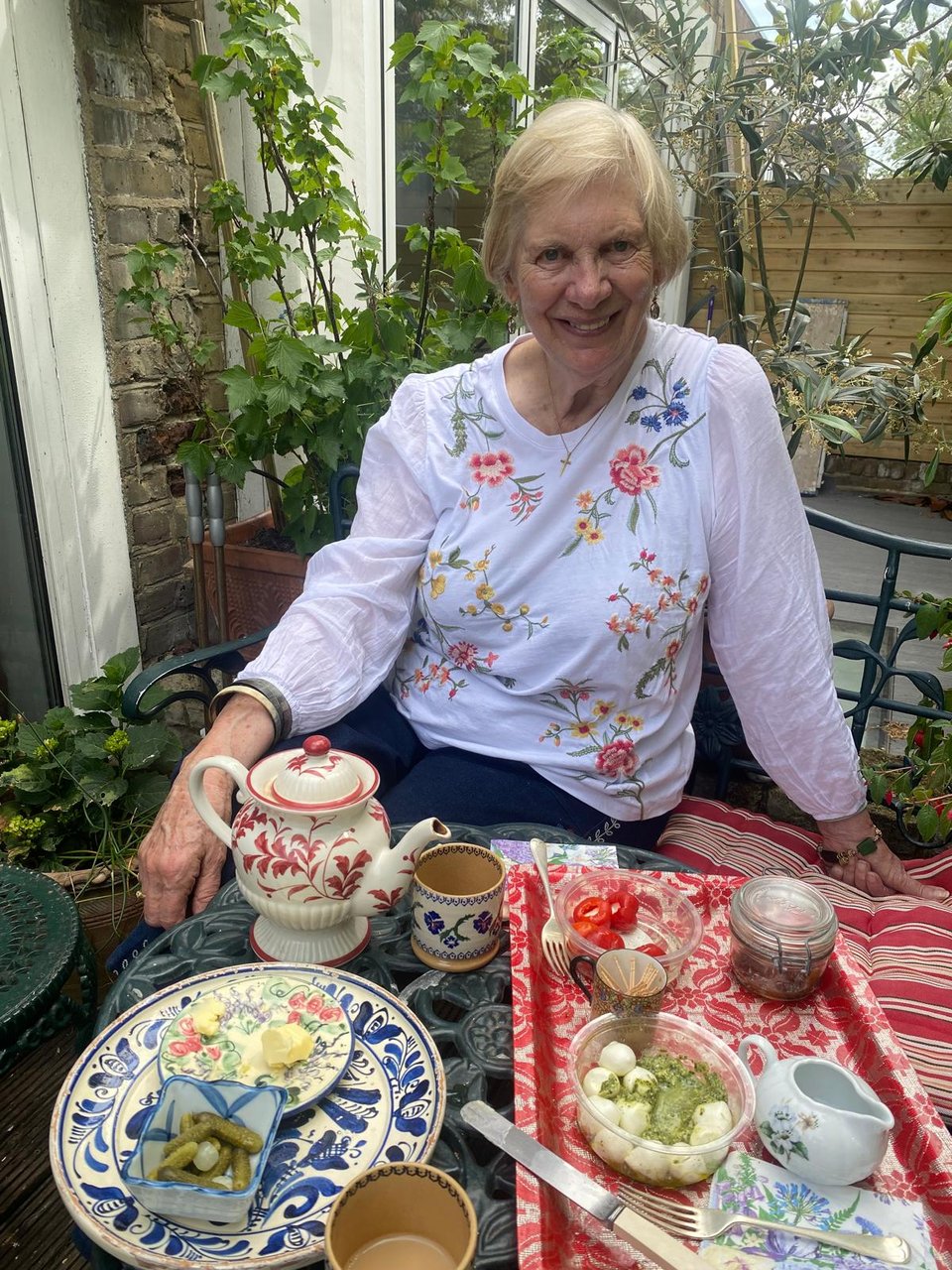 A woman sitting at a wrought-iron table smiles at the camera. The table is covered with small plates of food and a teapot and two cups.