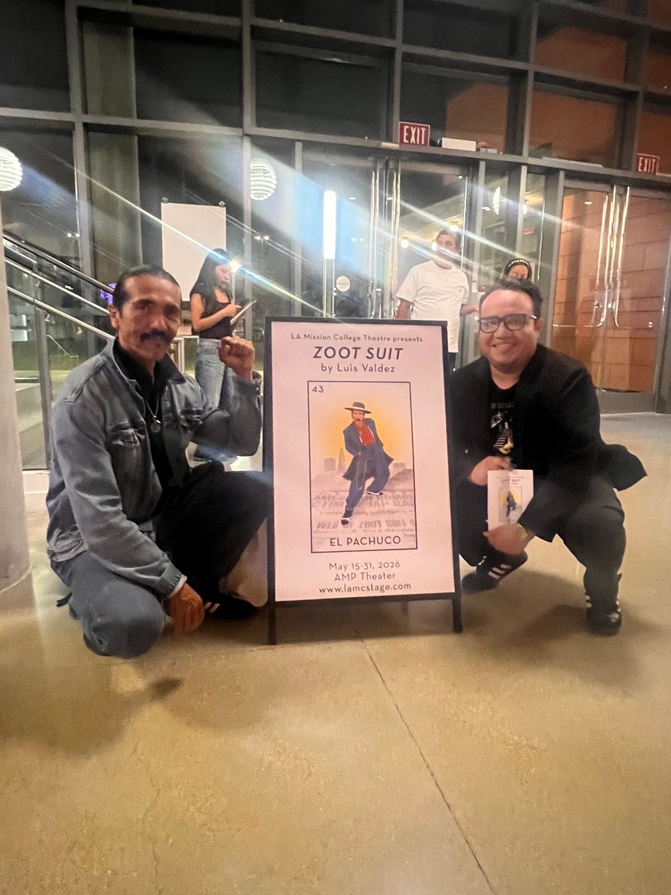 Raúl Cardona and Henry Barajas doing the cholo squat near a fold out banner to promote the Zoot suit play in the lobby of the theater.