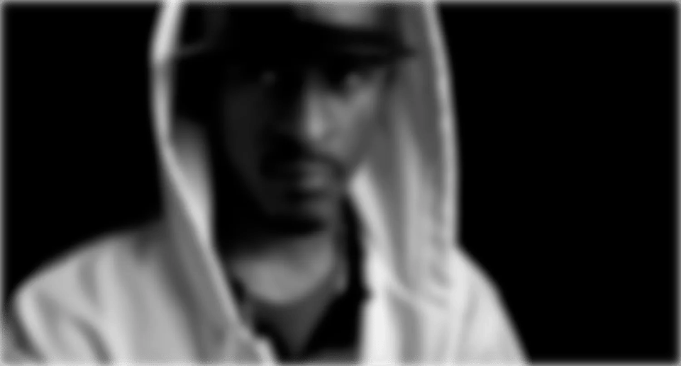 A blurred image of Rakim in a dark cap and white hoodie