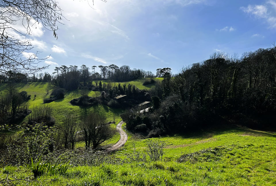 A undulating parkland on a bright morning. There are trees along the ridges and steep slopes. A track emerges from one side, and leads up to where several slate roofs are almost hidden in the trees.