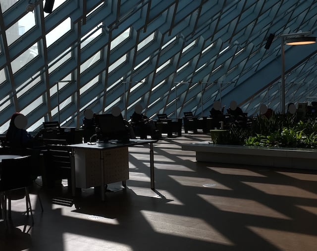 A view of people sitting at desks and in chairs under the slanting wall/ceiling of the Seattle Public Library Central Library. The slanting wall/ceiling is made up of glass panes in the shape of diamonds delineated by metal beams. The diamond pattern shows up on the wooden floor as sunlight streams through.