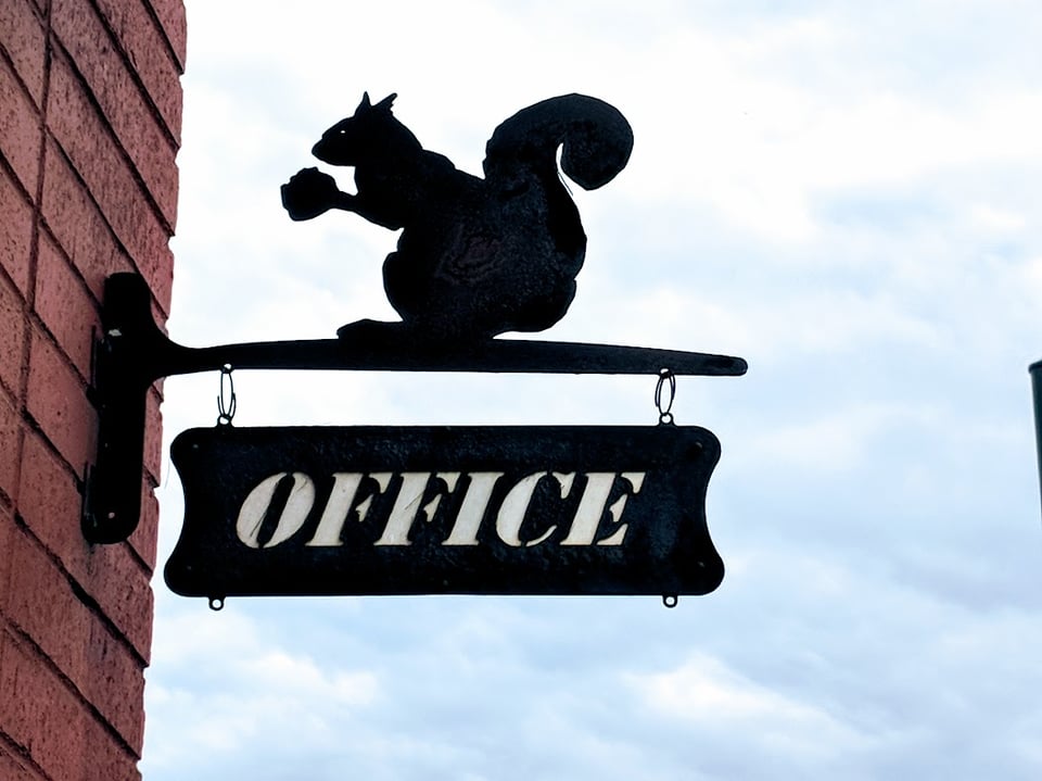It's a brick wall with a sign bolted to it. The sign says OFFICE in a slanty font, and there's a metal squirrel grabbing a nut on top.