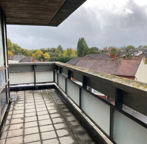 A balcony that runs around a building. It has a concrete and glass side, over which house rooftops and trees are visible. The blacony is paved with small square cream tiles, which are looking distinctly overgrown with muck.