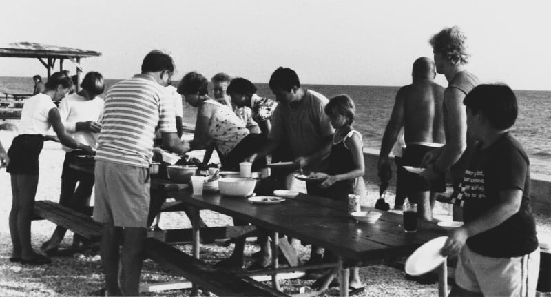 A group of people are getting plates of food around two picnic tables on the beach of the Gulf of Mexico.