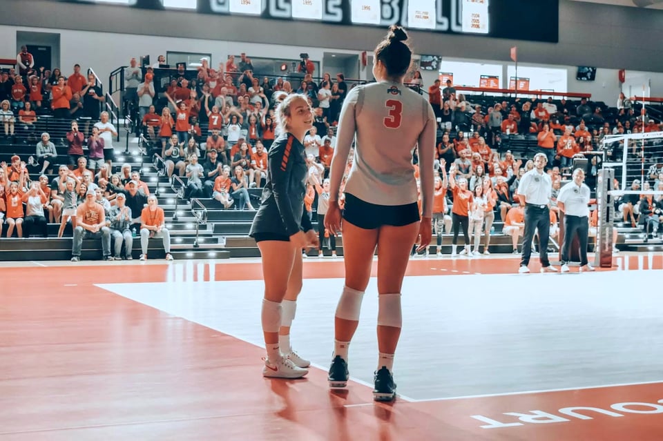 Mia Grunze stands with her back to the camera during a quiet moment in a volleyball match. She talks with another young woman who only comes up to Mia's shoulder, if that.