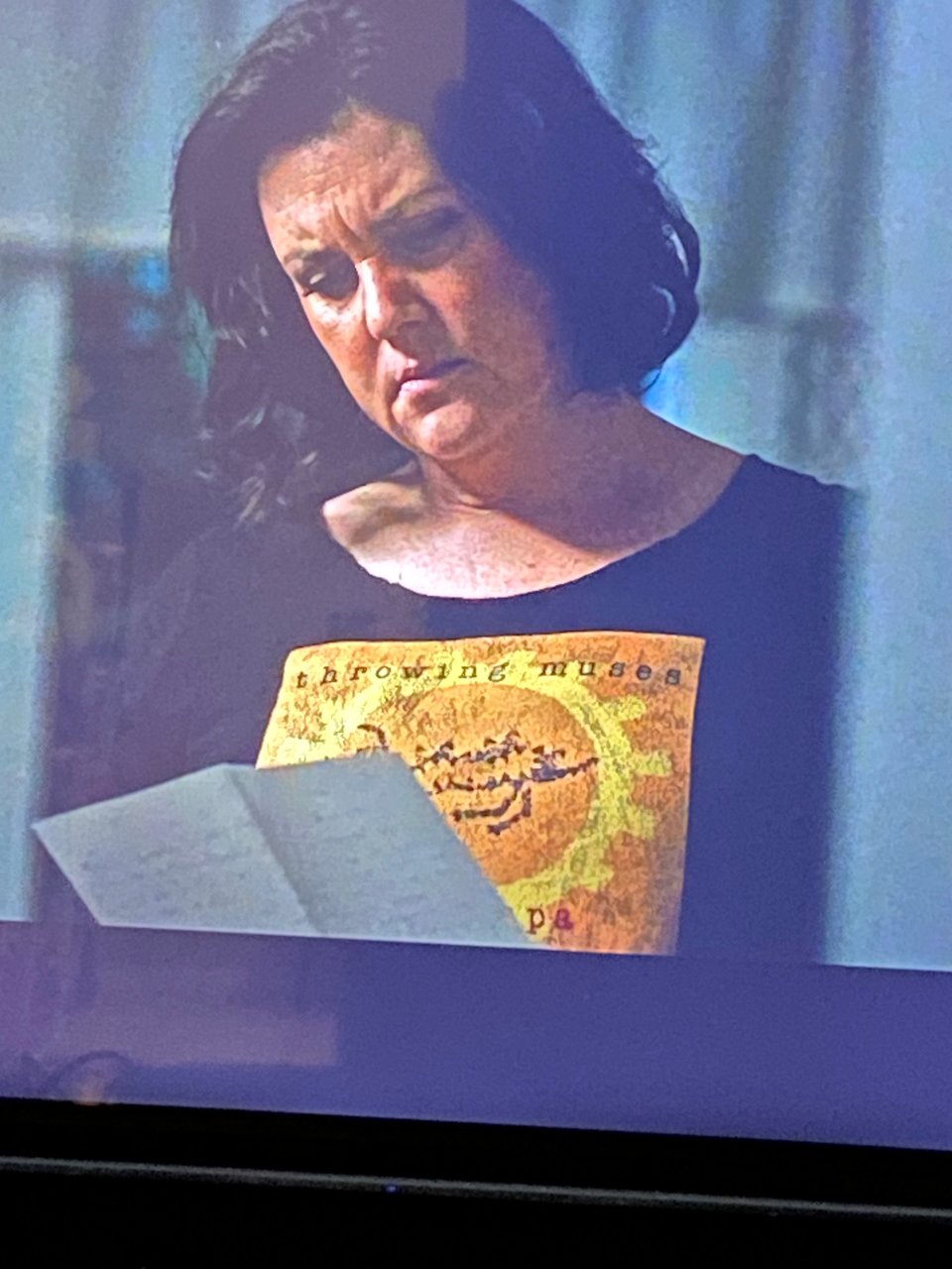 A picture of a TV with a shot from YELLOWJACKETS, with Melanie Lynskey, a woman in her late 40s, with black hair, in deep thought while reading a letter. She is wearing a black T Shirt with the cover of HUNKPAPA on it. The cover of HUNKPAPA: A dark yellow background. In the middle of it, taking most of the space, is a bright yellow circle with squares attached to the outside of it, suggesting a sun. Within it is a drawing of a creature, done in a kind of Paleolithic cave style. It has four legs, horns, ridges on its back, a tail.