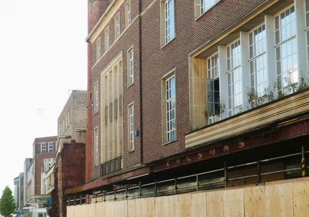 A 1950s department store in Exeter. It is brick clad, with stone window enclosures and a narrow balcony. The exposed girders above the builders' hoardings are red, with the name colson's picked out in yellow paint.