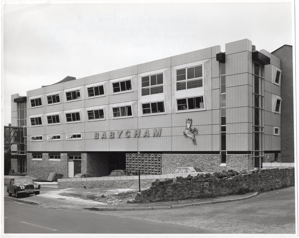 A rectangular office block. The ground floor is rubblestone, then there are three storeys that are clad in vertical rectangular tiles. The top two stories have windows set into cast concrete hoods. Along the side is a babysham sign in a bold blocky font. And the leaping fawn Babycham icon.