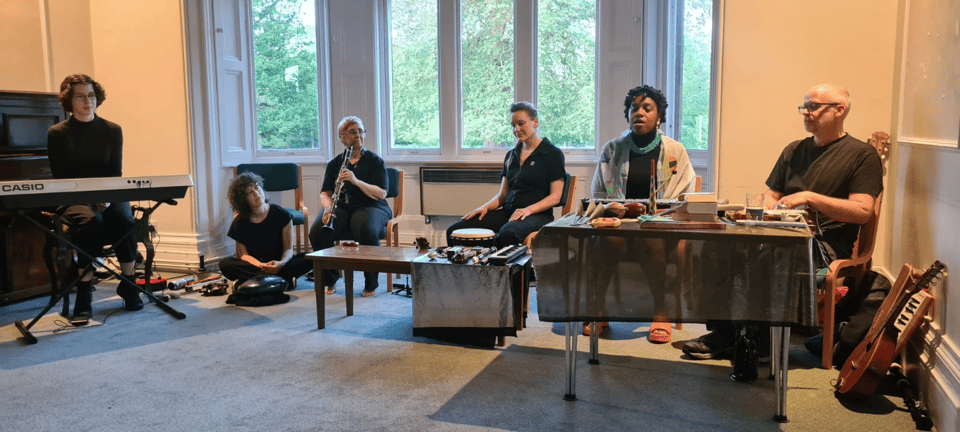 A group of musicians performs in a room with large windows, featuring a variety of instruments including a keyboard, flute, and percussion.