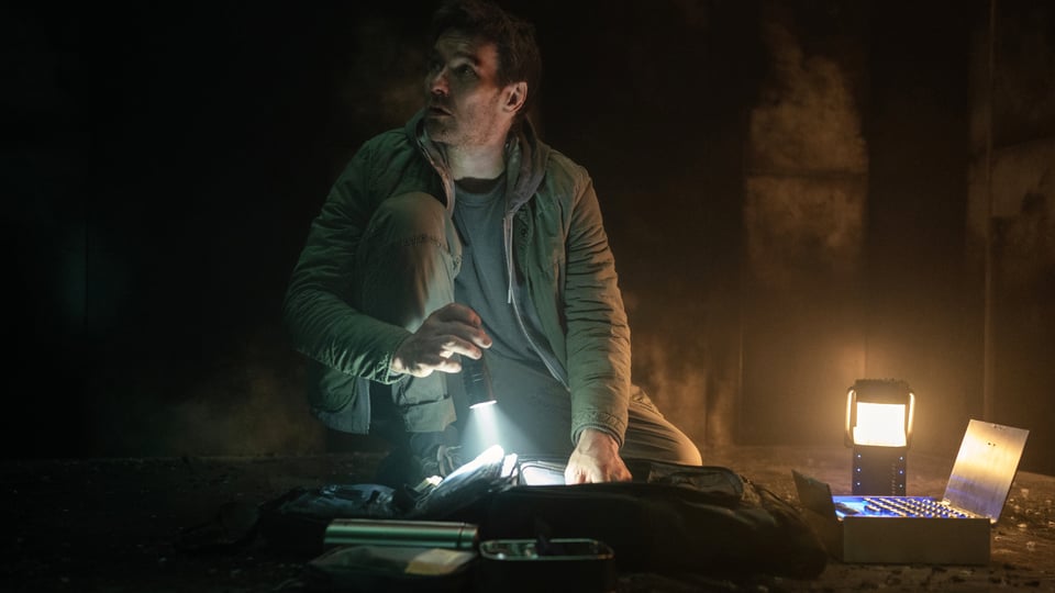 A still from the Apple TV+ show, Dark Matter. Joel Edgerton is kneeling in a dark corridor with a flashlight and a lantern.