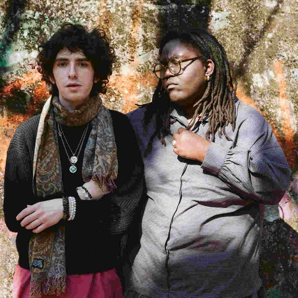 A man with unruly dark hair wearing a paisley scarf over a black sweater and red pants stands to the left of a woman with dreadlocks and glasses in a great jacket, who has her right arm around the man's back.