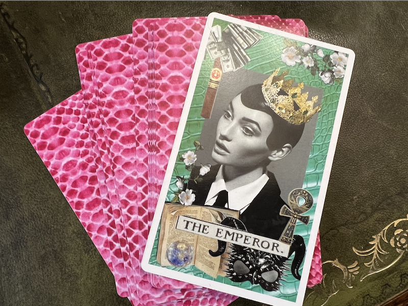 Top down photo of an Emperor tarot card sitting face up on a fanned out pile of facedown cards. The cards are laid on a vintage leather topped desk.