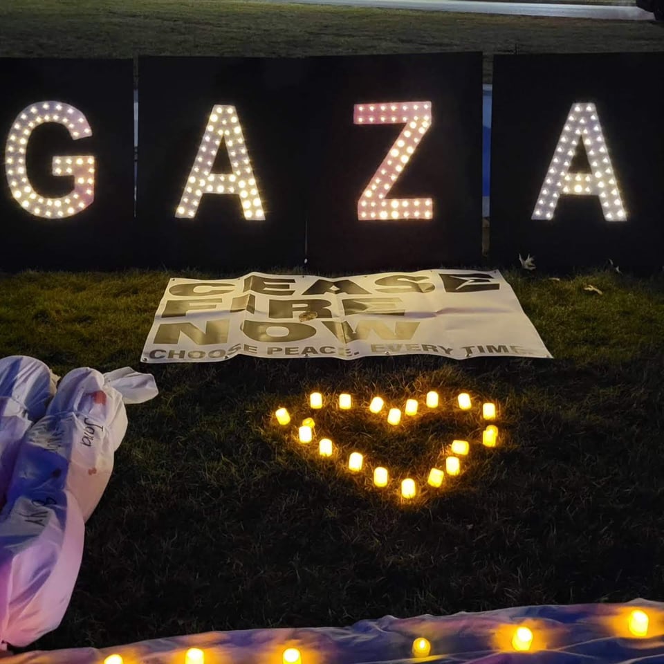 Black blocks with white letters and small LED lights spell out GAZA in the top of the frame. Below the letters is a white sign with black letters, "Ceasefire Now." Below that sign in the middle of the frame is an arrangement of tea light candles in a heart shape, and to the left edge of the photo are small white sheets representing the bodies of dead children wrapped for burial in Palestine.