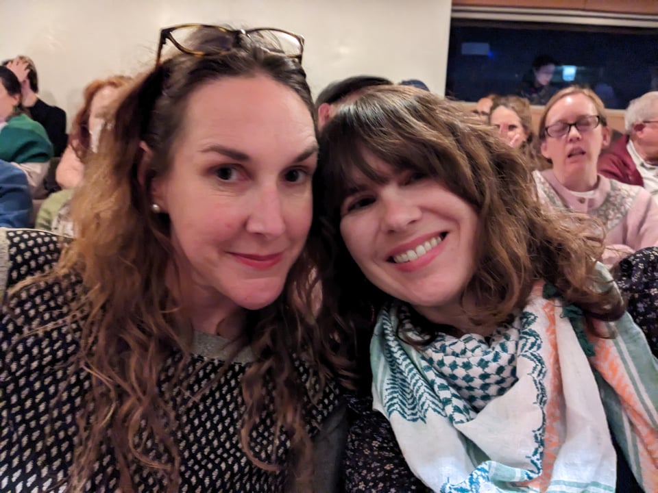 A smiling selfie of Summer and Lauren sitting in the theatre audience. Summer is wearing a black and white checked sweater. Lauren is wearing a black floral blouse and an orange, white and green Saoirse keffiyeh from Hirbawi.
