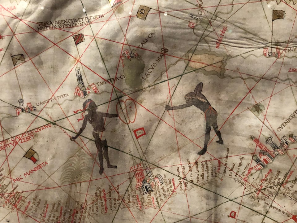 Medieval map detail showing two Black figures facing each other, one holding up a shield, the other aiming a bow and arrow. There are dainty little sketches of cities on the coast, which is bristling with place-names.