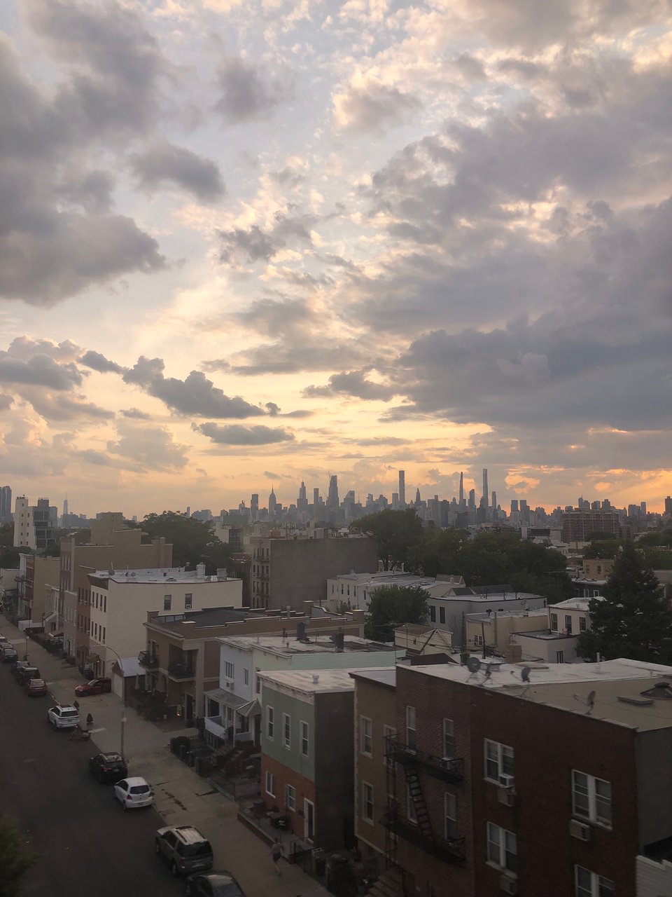 a photo of New York, showing a lo of lower-rise housing in front the silhouetted skyline of Manhattan. above, the sky is dramatically lit and cloudy.